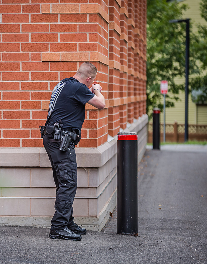 A police officer stands cautiously at the corner of a brick building during a training simulation.