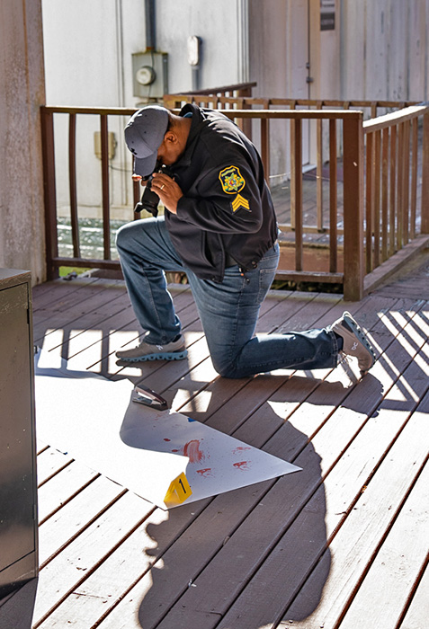 An officer with a camera takes a crime scene photo on a wooden deck.