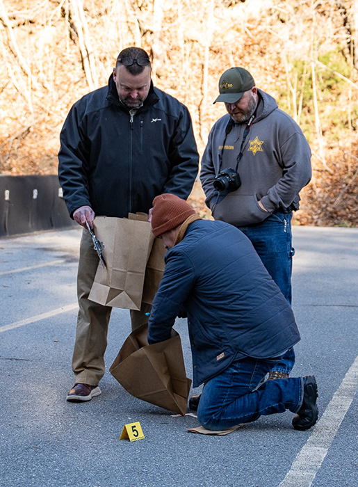 A group of three police officers stand on asphalt collecting evidence in paper bags.