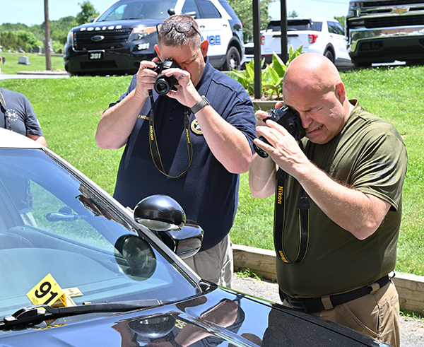 Two police officers practice crime scene photography techniques outdoors on a staged crime scene.
