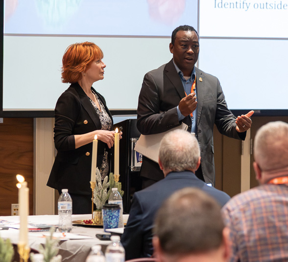 A woman and man stand at the front of a seated crowd during a communications symposium.