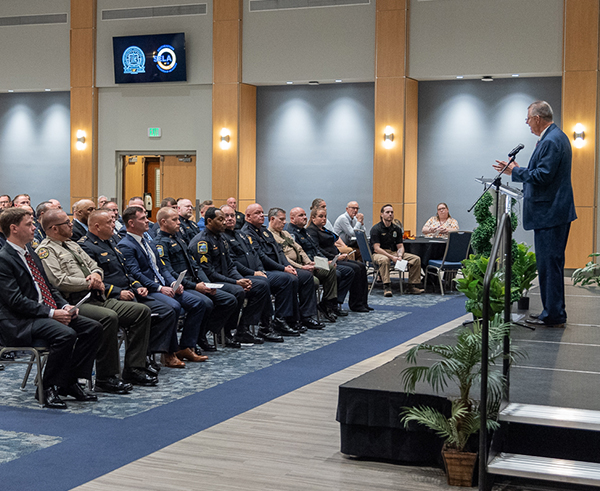 A group of police officers sit in a large group at a graduation ceremony and listen to a speaker onstage.