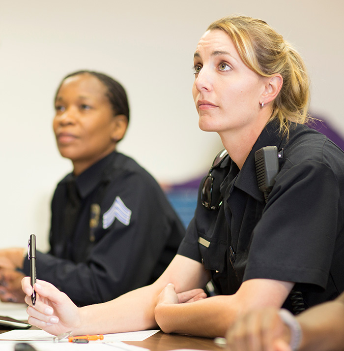 Two female police officers sit at a table and listen to a training seminar.