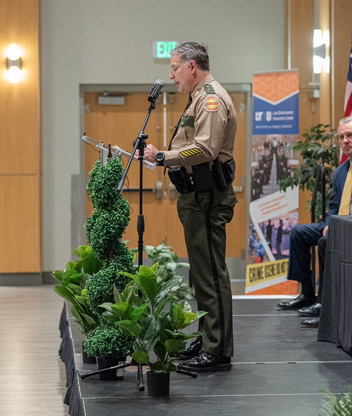 An officer in uniform gives a speech at a podium in a large room.
