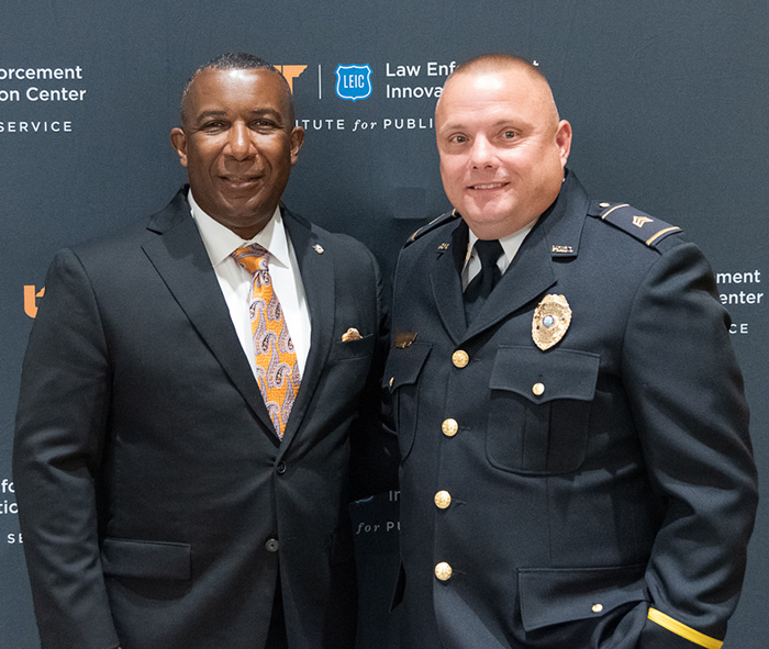 A man in a suit poses for a photo with a police officer in front of a dark backdrop.